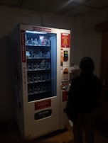 A dimly lit vending machine with various compartments, some containing products like snacks. A person stands in front of the machine, silhouetted in the darkness. The machine has instructions and pricing information displayed in different languages.