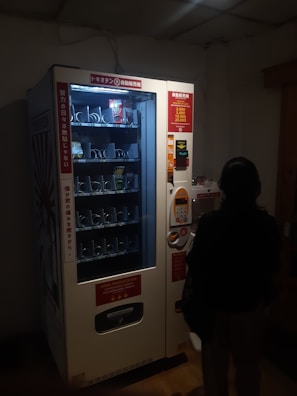 A dimly lit vending machine with various compartments, some containing products like snacks. A person stands in front of the machine, silhouetted in the darkness. The machine has instructions and pricing information displayed in different languages.