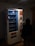 A dimly lit vending machine with various compartments, some containing products like snacks. A person stands in front of the machine, silhouetted in the darkness. The machine has instructions and pricing information displayed in different languages.