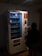 A dimly lit vending machine with various compartments, some containing products like snacks. A person stands in front of the machine, silhouetted in the darkness. The machine has instructions and pricing information displayed in different languages.