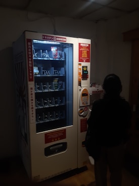 A dimly lit vending machine with various compartments, some containing products like snacks. A person stands in front of the machine, silhouetted in the darkness. The machine has instructions and pricing information displayed in different languages.