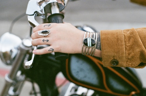 A hand with a tattoo of arrows is gripping the handlebar of a motorcycle. The person is wearing a brown corduroy jacket and has a large silver ring with a black stone and a silver bracelet with black accents. The motorcycle handles are shiny and metallic.