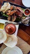 A meal featuring a variety of seafood arranged on a black rectangular plate with a banana leaf as a base. There are large shrimp, crab, and shellfish, accompanied by a tangy red sauce. A separate white dish holds a small brown clay pot and two sauces, one yellow and one brown. Alongside are fresh onion slices and lime on a separate tray with metal tongs. In the foreground, there are two white appam breads on a woven mat.