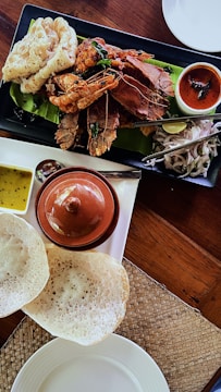 A meal featuring a variety of seafood arranged on a black rectangular plate with a banana leaf as a base. There are large shrimp, crab, and shellfish, accompanied by a tangy red sauce. A separate white dish holds a small brown clay pot and two sauces, one yellow and one brown. Alongside are fresh onion slices and lime on a separate tray with metal tongs. In the foreground, there are two white appam breads on a woven mat.