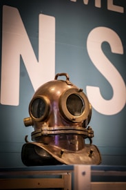 A vintage copper and brass diving helmet with round glass windows, resting on a wooden shelf against a teal background with partial white letters visible. The helmet has a tarnished patina, indicating age and use.