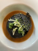 A gourmet dish featuring a dark, possibly black sesame or charcoal crisp, topped with fresh green herbs. It rests on a rich, brown sauce within a white plate, creating a striking visual contrast.
