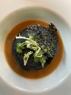 A gourmet dish featuring a dark, possibly black sesame or charcoal crisp, topped with fresh green herbs. It rests on a rich, brown sauce within a white plate, creating a striking visual contrast.