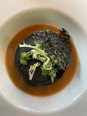 A gourmet dish featuring a dark, possibly black sesame or charcoal crisp, topped with fresh green herbs. It rests on a rich, brown sauce within a white plate, creating a striking visual contrast.