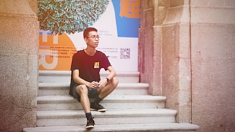 A person wearing a black t-shirt and glasses is sitting on stone steps in front of a colorful, abstract background featuring blue, orange, and white elements. The background includes text and a QR code.