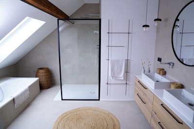 A contemporary bathroom with a minimalist design features a glass-enclosed shower with a black frame. Adjacent to the shower is a white bathtub, under a slanted ceiling with a skylight. The bathroom also includes a wooden storage basket, a round woven rug, and a double sink vanity with wooden drawers. Above the sinks, a large round mirror is installed, and two pendant lights hang from the ceiling. On the wall beside the sink, there is a metal rack with towels.