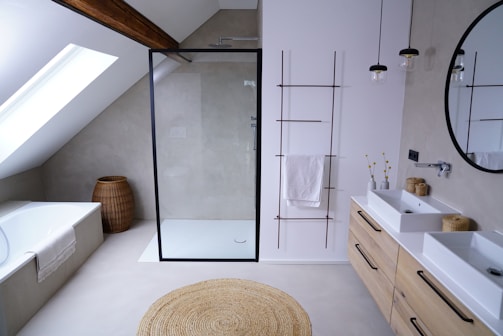 A contemporary bathroom with a minimalist design features a glass-enclosed shower with a black frame. Adjacent to the shower is a white bathtub, under a slanted ceiling with a skylight. The bathroom also includes a wooden storage basket, a round woven rug, and a double sink vanity with wooden drawers. Above the sinks, a large round mirror is installed, and two pendant lights hang from the ceiling. On the wall beside the sink, there is a metal rack with towels.
