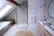 A contemporary bathroom with a minimalist design features a glass-enclosed shower with a black frame. Adjacent to the shower is a white bathtub, under a slanted ceiling with a skylight. The bathroom also includes a wooden storage basket, a round woven rug, and a double sink vanity with wooden drawers. Above the sinks, a large round mirror is installed, and two pendant lights hang from the ceiling. On the wall beside the sink, there is a metal rack with towels.