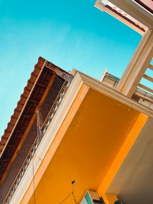 The image features an upward perspective of a building with colorful elements. The structure displays a bright orange wall that contrasts with a clear blue sky. There is a traditional-style roof with tiles extending over the top, and a balcony made of a wooden railing.