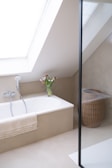 A minimalist bathroom with a large skylight providing natural light. The bathtub is integrated into a beige alcove, featuring a handheld showerhead and a fringed towel draped over the side. A clear vase with fresh tulips is placed at the corner of the tub. To the right, a woven wicker laundry basket sits on the floor. The space is enclosed by a glass partition, suggesting a shower area.