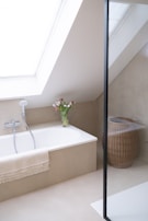 A minimalist bathroom with a large skylight providing natural light. The bathtub is integrated into a beige alcove, featuring a handheld showerhead and a fringed towel draped over the side. A clear vase with fresh tulips is placed at the corner of the tub. To the right, a woven wicker laundry basket sits on the floor. The space is enclosed by a glass partition, suggesting a shower area.
