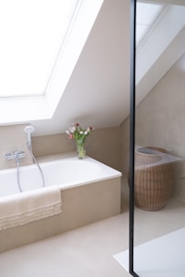 A minimalist bathroom with a large skylight providing natural light. The bathtub is integrated into a beige alcove, featuring a handheld showerhead and a fringed towel draped over the side. A clear vase with fresh tulips is placed at the corner of the tub. To the right, a woven wicker laundry basket sits on the floor. The space is enclosed by a glass partition, suggesting a shower area.