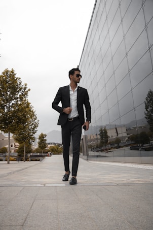 A man dressed in a sleek black suit and white shirt walks confidently down a paved urban pathway, flanked by modern architecture with reflective glass panels and neatly arranged trees. The sky appears overcast, adding a sophisticated tone to the environment.