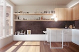 A modern kitchen with minimalist design featuring wooden cabinets and a center island with two white bar stools. Floating shelves are adorned with books, small plants, and decorative items. Natural light streams through a large window, enhancing the earthy and neutral color palette. Pendant lights hang above the island, adding a warm ambiance.