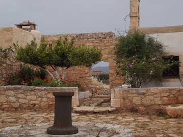 An old stone courtyard with a rustic aesthetic, featuring a central pedestal or well surrounded by stone walls and an archway. Lush greenery and vibrant flowers add color to the scene, with a clear view of the countryside beyond. There is an old stone structure in the background, and a cloudy sky overhead.