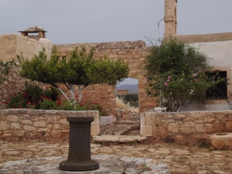 An old stone courtyard with a rustic aesthetic, featuring a central pedestal or well surrounded by stone walls and an archway. Lush greenery and vibrant flowers add color to the scene, with a clear view of the countryside beyond. There is an old stone structure in the background, and a cloudy sky overhead.