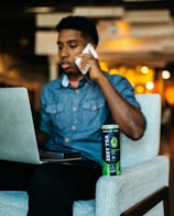 A person is sitting on a chair in an indoor setting, holding a phone to their ear and looking at a laptop screen. A can of Zest Tea energy drink is placed on the armrest of the chair. The background is softly blurred with warm lighting.