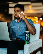 A person is sitting on a chair in an indoor setting, holding a phone to their ear and looking at a laptop screen. A can of Zest Tea energy drink is placed on the armrest of the chair. The background is softly blurred with warm lighting.