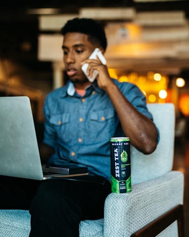 A person is sitting on a chair in an indoor setting, holding a phone to their ear and looking at a laptop screen. A can of Zest Tea energy drink is placed on the armrest of the chair. The background is softly blurred with warm lighting.