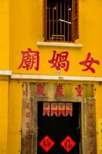 A yellow building with a window featuring metal bars above a doorway. Red Chinese characters are prominently displayed on the walls and above the door. The entrance is adorned with traditional red banners featuring calligraphy.