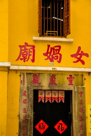 A yellow building with a window featuring metal bars above a doorway. Red Chinese characters are prominently displayed on the walls and above the door. The entrance is adorned with traditional red banners featuring calligraphy.