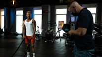 A man in a white tank top and red shorts is exercising with battle ropes in a gym. Another person wearing a black shirt and jeans is filming or taking pictures with a camera, while wearing a mask. The gym is equipped with exercise machines and the lighting is dim, coming from outside through large windows.