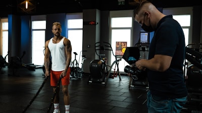 A man in a white tank top and red shorts is exercising with battle ropes in a gym. Another person wearing a black shirt and jeans is filming or taking pictures with a camera, while wearing a mask. The gym is equipped with exercise machines and the lighting is dim, coming from outside through large windows.