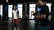 A man in a white tank top and red shorts is exercising with battle ropes in a gym. Another person wearing a black shirt and jeans is filming or taking pictures with a camera, while wearing a mask. The gym is equipped with exercise machines and the lighting is dim, coming from outside through large windows.