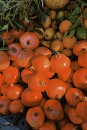 A pile of pomegranates and other fruits are surrounded by green leaves. The fruits are stacked closely together, showcasing their round shapes and vibrant colors.