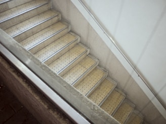 A stairway with beige, perforated steps bordered by metallic strips, enclosed by a concrete wall. There is also a metal handrail on the right side and a partial view of a glass barrier on the left.