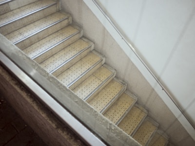 A stairway with beige, perforated steps bordered by metallic strips, enclosed by a concrete wall. There is also a metal handrail on the right side and a partial view of a glass barrier on the left.