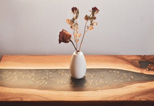 A white ceramic vase holds a few dried roses with brown and red petals and leaves. The vase is placed on a wooden table that has a smooth, flowing design.