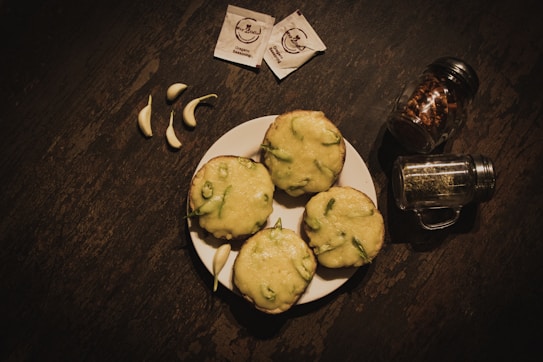 A plate with four slices of bread topped with melted cheese and green pepper is placed on a dark rustic surface. Surrounding the plate are garlic cloves, seasoning packets, and two spice jars containing red pepper flakes and dried herbs.