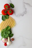 A flat lay arrangement of fresh tomatoes, spinach leaves, garlic, pasta sheets, and a single cherry tomato on a white marbled surface.
