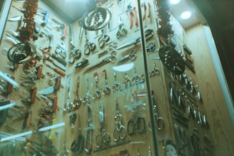 A display cabinet filled with a variety of scissors and cutting tools. The tools are arranged neatly against a wooden backdrop and include different types of scissors, knives, and multi-tools. The scene is decorated with Christmas-themed ornaments, such as tinsel and metallic decorations hanging from above.