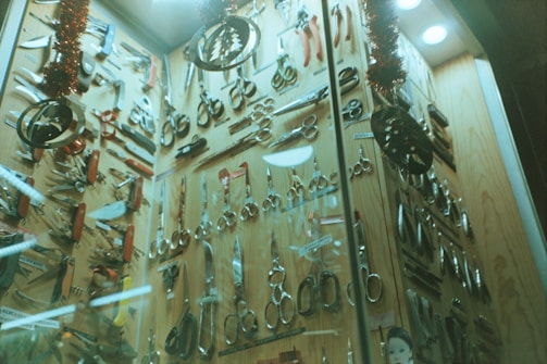 A display cabinet filled with a variety of scissors and cutting tools. The tools are arranged neatly against a wooden backdrop and include different types of scissors, knives, and multi-tools. The scene is decorated with Christmas-themed ornaments, such as tinsel and metallic decorations hanging from above.