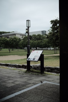 A park setting with a paved walkway leading to an informational sign or display board. The sign is positioned on a pair of black stands and features text and images. Surrounding the area are green grassy fields, trees, and a modern building in the background. The sky is overcast, contributing to a subdued atmosphere.