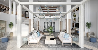 A spacious, modern living area with high ceilings and an open layout. The room features white and light grey tones with wooden accents. There are two couches facing each other with a large rectangular coffee table between them. The space is decorated with blue and grey cushions, a floor rug, and plants. Multiple pendant lamps hang from the ceiling. The room has a second level with railings and additional seating areas, and artwork is displayed on the walls.