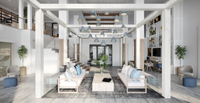 A spacious, modern living area with high ceilings and an open layout. The room features white and light grey tones with wooden accents. There are two couches facing each other with a large rectangular coffee table between them. The space is decorated with blue and grey cushions, a floor rug, and plants. Multiple pendant lamps hang from the ceiling. The room has a second level with railings and additional seating areas, and artwork is displayed on the walls.