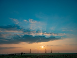 A landscape with wind turbines silhouetted against a bright sunset, casting rays through scattered clouds. The foreground features a green field, while the sky transitions from deep blue to warm orange.