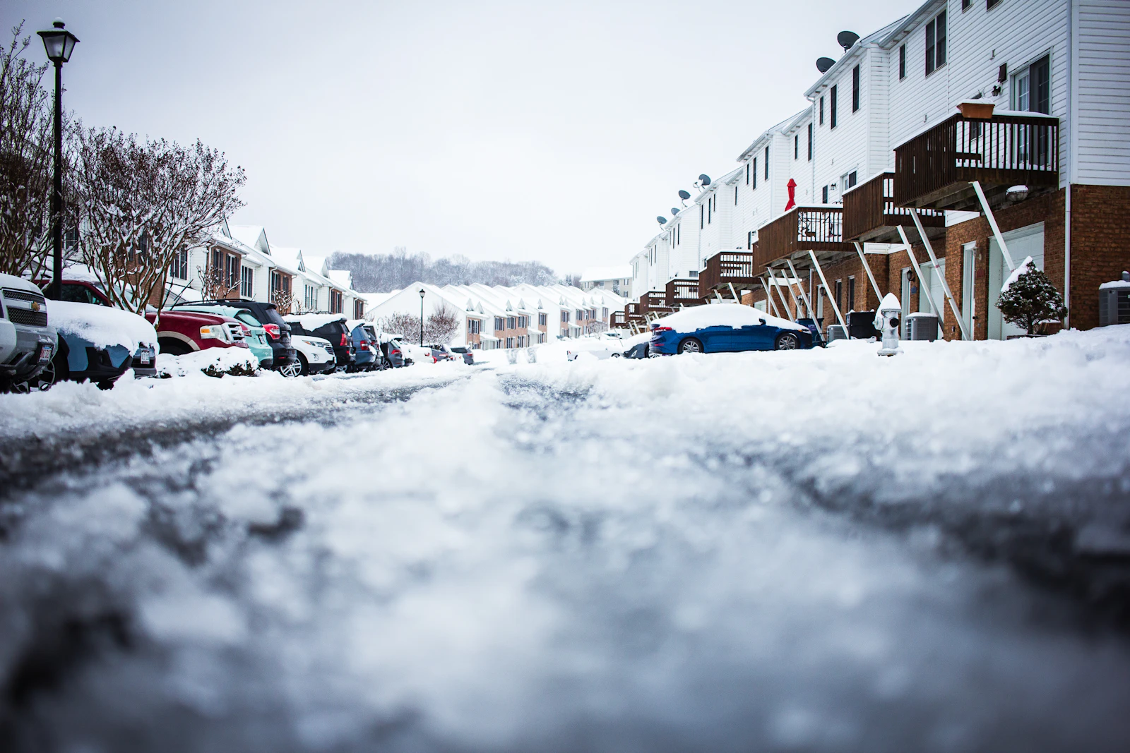 A snow-covered residential street lined with houses and parked cars on a winter day
