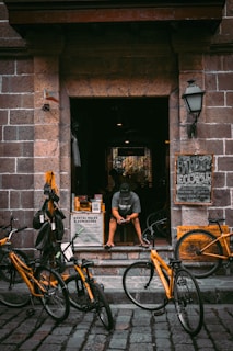 A person sits on the steps of a shop, looking at their phone. Several bicycles with bright yellow frames are parked in front of the entrance. The shop is constructed from stone and has a rustic charm. Signs around the entrance advertise bicycle rentals.