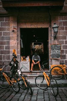 A person sits on the steps of a shop, looking at their phone. Several bicycles with bright yellow frames are parked in front of the entrance. The shop is constructed from stone and has a rustic charm. Signs around the entrance advertise bicycle rentals.