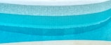 A close-up view of a curved swimming pool edge with a mosaic tile pattern. The tiles are primarily blue with varying shades, creating a gradient effect in the water. The upper portion of the pool is lined with white tiles, and the pool water appears clean and turquoise.