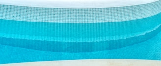 A close-up view of a curved swimming pool edge with a mosaic tile pattern. The tiles are primarily blue with varying shades, creating a gradient effect in the water. The upper portion of the pool is lined with white tiles, and the pool water appears clean and turquoise.