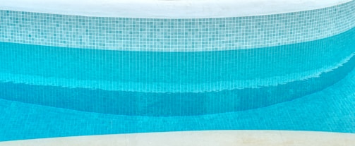 A close-up view of a curved swimming pool edge with a mosaic tile pattern. The tiles are primarily blue with varying shades, creating a gradient effect in the water. The upper portion of the pool is lined with white tiles, and the pool water appears clean and turquoise.